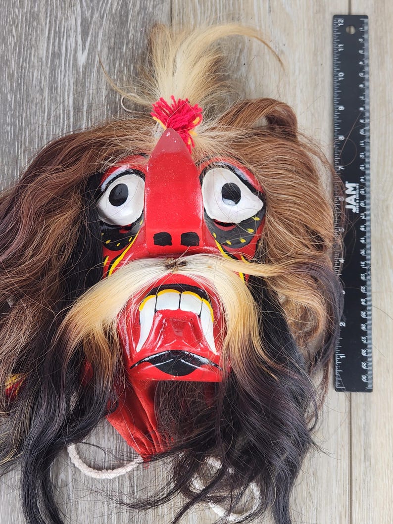 Pu&ograve; includere: Una maschera artigianale con un'espressione feroce. La maschera presenta un volto rosso con occhi bianchi, un naso prominente e una bocca ampia che rivela denti bianchi. &Egrave; adornata da capelli castani, neri e biondi e da un nappa rossa. La maschera &egrave; probabilmente utilizzata per scopi culturali o teatrali.