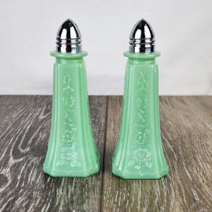 May include: Two vintage, light green salt and pepper shakers with chrome tops. The glass shakers have a floral and leaf design. The shakers are displayed on a wooden surface.