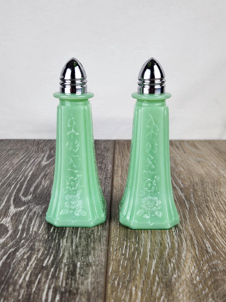 May include: Two vintage, light green salt and pepper shakers with chrome tops. The glass shakers have a floral and leaf design. The shakers are displayed on a wooden surface.