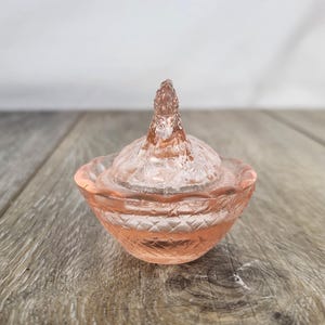 May include: A small, pink glass trinket dish with a lid. The dish has a diamond pattern around the base and a textured lid with a pointed finial. The glass has a soft, translucent appearance. The dish is likely vintage or antique.