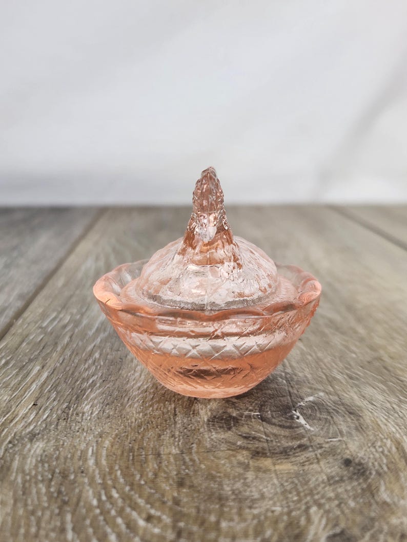 May include: A small, pink glass trinket dish with a lid. The dish has a diamond pattern around the base and a textured lid with a pointed finial. The glass has a soft, translucent appearance. The dish is likely vintage or antique.