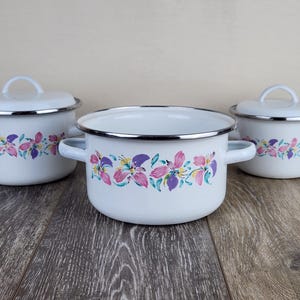 May include: Three white enamel pots with lids and handles. Each pot features a floral design in pink, purple, and yellow. The pots are arranged on a wooden surface.