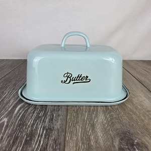 May include: A light blue rectangular butter dish with a matching lid featuring a curved handle. The word Butter is printed in black script lettering on the front of the lid. The dish has a black rim around the base and a glossy enamel finish.