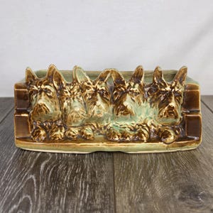 May include: A rectangular ceramic planter featuring a relief design of five Scottish Terrier dog heads across the front. The piece has a glossy finish with a color palette of deep brown on the dogs and a light sage green glaze on the background.