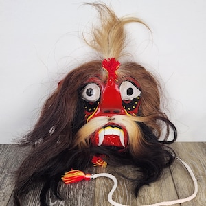 Pu&ograve; includere: Maschera decorativa fatta a mano con un'espressione feroce. La maschera presenta un volto rosso con denti bianchi, lunghi baffi bianchi e capelli scuri. Un cordino bianco con nappa pende dalla maschera.
