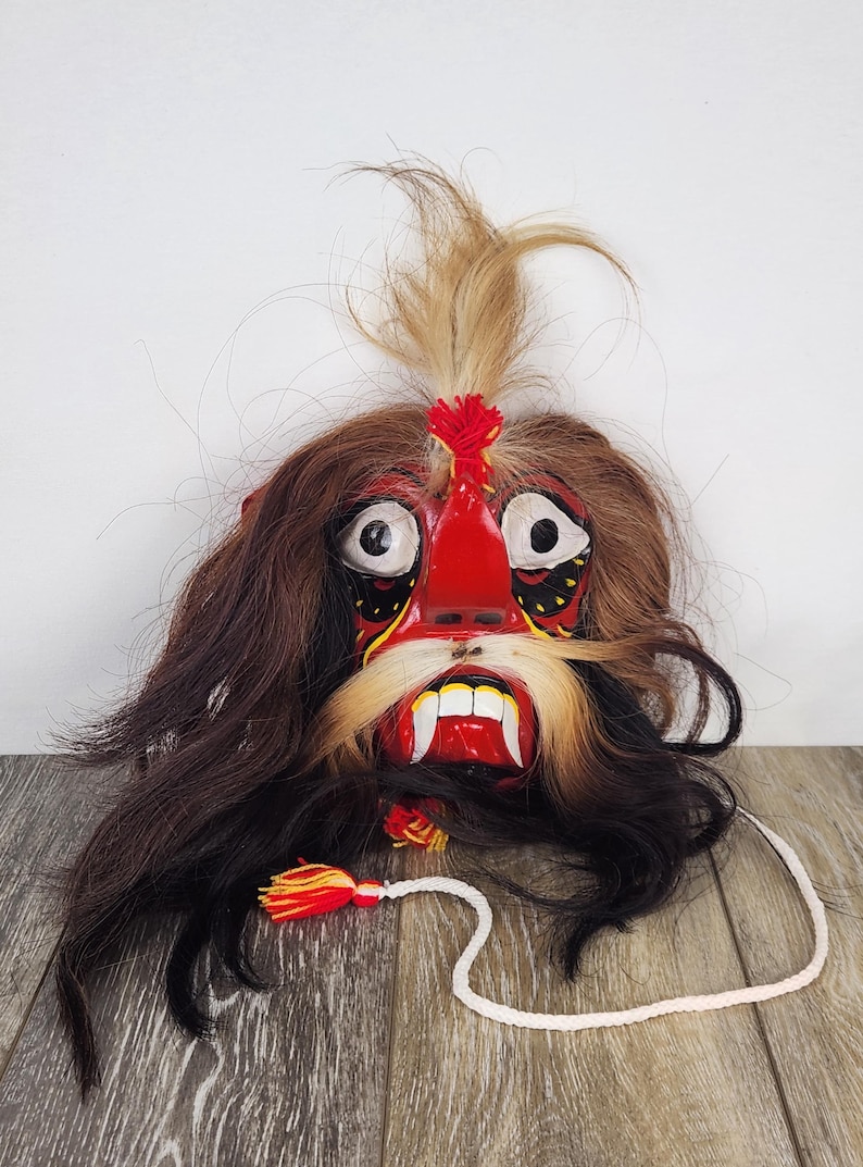 Pu&ograve; includere: Maschera decorativa fatta a mano con un'espressione feroce. La maschera presenta un volto rosso con denti bianchi, lunghi baffi bianchi e capelli scuri. Un cordino bianco con nappa pende dalla maschera.