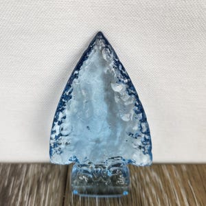 May include: A small, light blue glass arrowhead sculpture with a textured surface. The arrowhead is on a clear glass base.