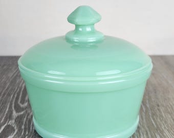 May include: A light mint green jadeite glass bowl with a matching lid. The bowl features a smooth, rounded body with a slightly tapered base. The lid has a prominent, faceted, hexagonal knob handle centered on top, providing a secure grip.