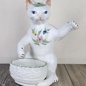 May include: A white ceramic cat figurine with a basket. The cat has blue eyes, a pink nose, and floral accents. The basket has a woven texture and a green rim. The figurine is a decorative item.