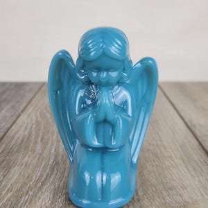 May include: A teal-colored angel figurine with folded hands in prayer. The angel has detailed wings and a serene expression. The figurine is made of a glossy material and sits on a wooden surface. A decorative religious item.