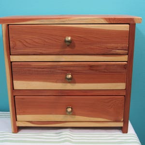 May include: A small wooden chest of drawers with three drawers. The chest is made of reddish-brown wood with lighter wood accents. Each drawer has a round, brass-coloured knob. The chest is sitting on a white and green striped surface.