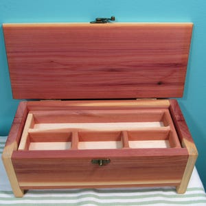 May include: Open wooden jewelry box with a hinged lid. The box is made of reddish-brown cedar wood with a light-colored interior and small compartments. A metal clasp secures the lid.