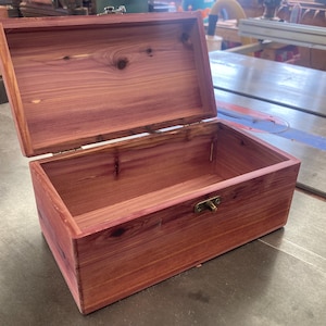 May include: A rectangular cedar wood box with a hinged lid and a brass-coloured metal latch. The box features a natural reddish-brown wood grain with visible knots and a smooth finish. The lid is open, revealing the interior of the wooden container.