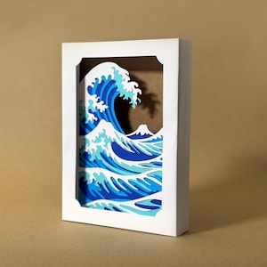 May include: A 3D paper art piece depicting the iconic Japanese wave, with shades of blue and white. The artwork is framed in a white box, showcasing the layered design and a glimpse of a mountain in the background. The piece is a decorative item.