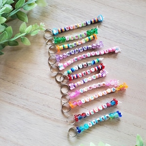 May include: A collection of eleven personalized keychains, each featuring a metal ring attached to a strand of colorful plastic beads. The beads spell out various names including Olivia, James, Noah, Harper, Charlotte, Liam, Levi, Sophia, Isabella, Lucas, and Ryder.