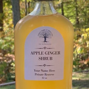 May include: A clear glass bottle with a black cap filled with a light yellow liquid. The bottle has a white label with the text "Forest Farms of Maryland", "Olde Thyme Fruits and Such", "Apple Ginger Shrub", "Your Name Here", "Private Reserve", and "12 oz".
