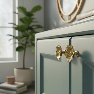 Peut inclure: Deux boutons de placard en métal doré en forme de fleurs ouvertes avec des pétales détaillés et un centre texturé. Les boutons sont fixés sur une porte de placard vert sauge mat avec un panneau encastré, une finition lisse et des bords nets.