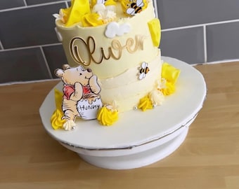 May include: A two-tiered yellow cake decorated with Winnie the Pooh, bees, and the name "Oliver" in gold script. The cake is topped with a number "1" and sits on a white cake stand. Yellow frosting and decorative elements complete the design.