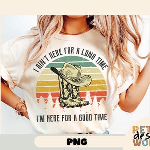 Puede incluir: Camiseta color crema con un diseño de sombrero y bota de vaquero sobre un fondo de puesta de sol. El texto dice: "I ain't here for a long time, I'm here for a good time."