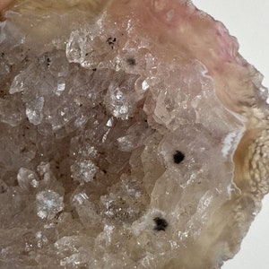 May include: A close-up of a geode, showcasing a cavity filled with sparkling, translucent crystals. The outer edge of the geode is a light beige color, with hints of pink and white. Two small black spots are visible within the crystal formation.