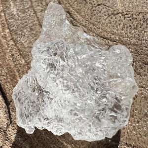 Può includere: Un cristallo trasparente di forma irregolare con una superficie ruvida e strutturata. Il cristallo è posizionato su una superficie di legno marrone invecchiato, con venature e crepe visibili. Le sfaccettature del cristallo catturano la luce, creando un effetto scintillante.