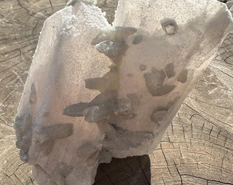 May include: A large, translucent quartz crystal cluster with a light, frosted appearance. The crystal has a rough, textured surface and is accented with smaller, pale green crystal formations. The quartz sits on a weathered, wooden surface.