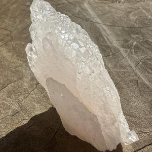Puede incluir: Un gran cristal de cuarzo transparente con un tono rosado. El cristal tiene un c&uacute;mulo de cristales m&aacute;s peque&ntilde;os en la parte superior y una base lisa y plana.