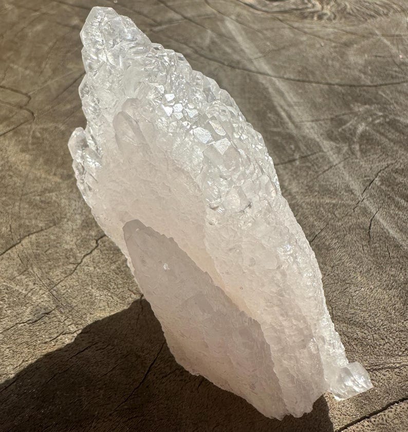 Puede incluir: Un gran cristal de cuarzo transparente con un tono rosado. El cristal tiene un c&uacute;mulo de cristales m&aacute;s peque&ntilde;os en la parte superior y una base lisa y plana.