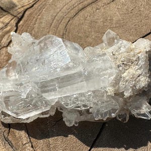 May include: A cluster of clear, crystalline minerals with a layered structure. The crystals are translucent and reflect light, showcasing their natural formations. The mineral formation is set against a textured, wooden surface.