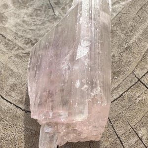 Puede incluir: Un gran cristal de selenita rosa claro, estriado y ligeramente transl&uacute;cido. El cristal descansa sobre una superficie de madera.