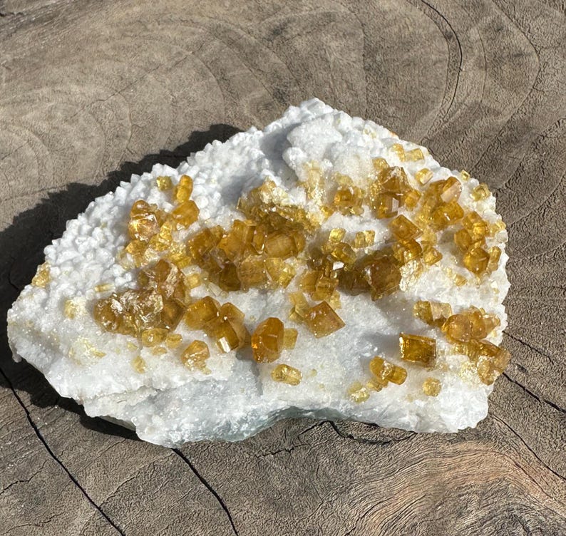 May include: A natural mineral specimen featuring a white matrix covered with clusters of golden-yellow crystals. The crystals have a sparkling, crystalline appearance, contrasting with the rough, textured base. The specimen is displayed on a wooden surface.