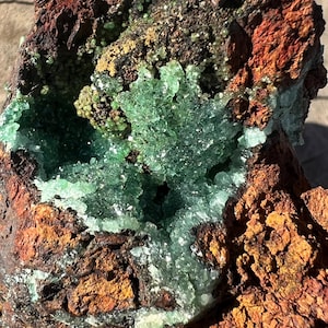 May include: A close-up of a green crystal geode embedded in a rough, rusty brown rock. The crystal is translucent and has a sparkly, iridescent quality.