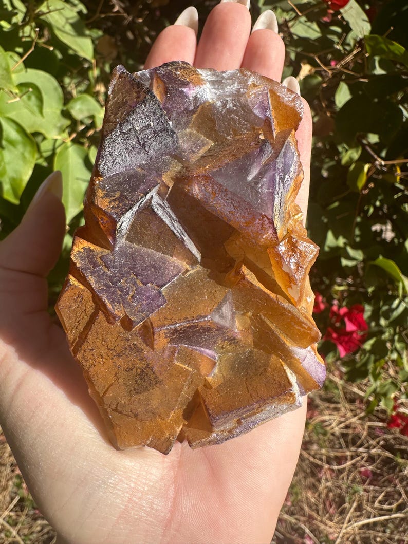 May include: A large, multi-faceted crystal cluster with a mix of purple and orange hues. The crystal has a rough, natural texture and is a beautiful example of a fluorite specimen.