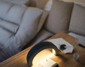 May include: A dark blue crescent-shaped lamp with a white light illuminates a small notebook on a wooden table. The lamp is on a bedside table next to a bed with white pillows and a beige throw pillow.