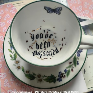 May include: A white teacup and saucer set with a green trim. The teacup has a butterfly and the words "you've been poisoned" in black. The saucer is decorated with floral patterns, creating a whimsical design.