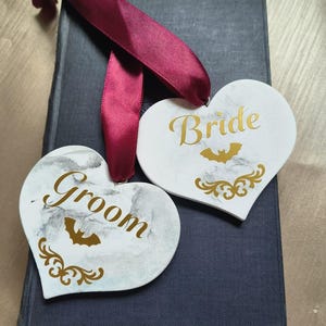 May include: Two heart-shaped wedding decorations, one reading "Groom" and the other "Bride" in gold script. Each heart has a bat and decorative flourishes. A burgundy ribbon is attached. The hearts rest on a dark blue book.
