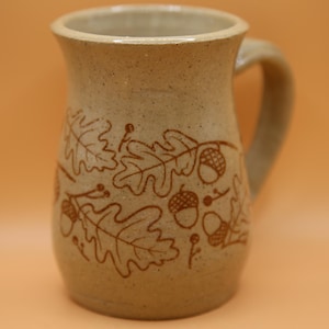 May include: A beige ceramic mug with a speckled finish and a handle on the right side. The exterior surface features a dark brown etched pattern of oak leaves and acorns wrapping around the body of the vessel in a repeating botanical design.