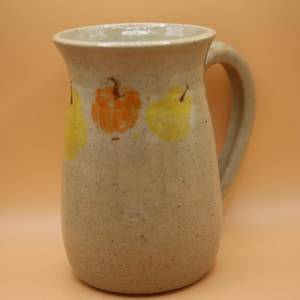 May include: A beige ceramic pitcher with a speckled texture and a single handle. The exterior features a hand-painted design of three small gourds in shades of orange and yellow arranged horizontally around the upper portion of the vessel.