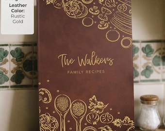 Op de afbeelding: Een donkerbruin receptenboek met gouden illustraties van groenten, kruiden en keukengerei. De kaft bevat de tekst "The Walkers" en "FAMILY RECIPES" in gouden letters. Een wit label aan het boek vermeldt "Design #: 1" en "Leather Color: Rustic Gold".