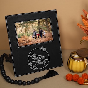 May include: Black leather photo frame with a white stitched border. The frame has a photo of a family in a forest and a personalized inscription that reads "The Walker Family".