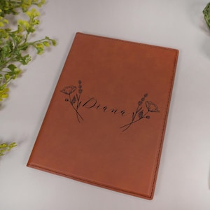 May include: A brown leather journal with the name "Diana" engraved on the front cover. The journal is decorated with two floral designs, each featuring a poppy flower and stems.