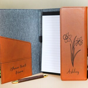 May include: A brown leatherette folder with a gray fabric exterior. The folder has a pocket on the front and a notepad inside. The front of the folder is personalized with the name "Ashley" and a floral design.