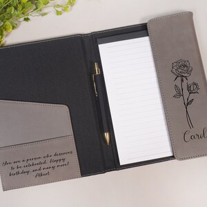 May include: A gray leatherette folder with a black interior and a pen holder. The folder has a pocket on the front with the text "You are a person who deserves to be celebrated. Happy birthday and many more! - Filbert". The back of the folder has a black and white illustration of a rose and the name "Carol" engraved on it.