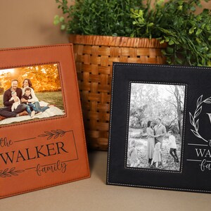 May include: Two leather photo frames with a black and brown color scheme. The frames have a stitched border and a personalized inscription that reads "the WALKER family". One frame features a color photo of a family of four, while the other frame features a black and white photo of a family of four.