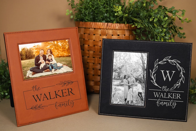 May include: Two leather photo frames with a black and brown color scheme. The frames have a stitched border and a personalized inscription that reads "the WALKER family". One frame features a color photo of a family of four, while the other frame features a black and white photo of a family of four.