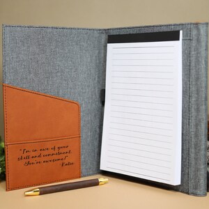 May include: A gray and brown leatherette journal with a notepad and a pen. The journal has a pocket on the front with the inscription "I'm in awe of your skill and commitment. You're awesome! Katie".