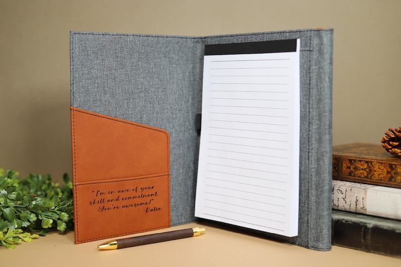 May include: A gray and brown leatherette journal with a notepad and a pen. The journal has a pocket on the front with the inscription "I'm in awe of your skill and commitment. You're awesome! Katie".