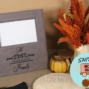 May include: A gray faux leather picture frame with a white mat and a black engraved inscription that reads "The Anderson Family".