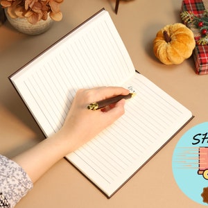 May include: A brown leather-bound journal with lined pages. A person is writing in the journal with a gold-tipped pen. The journal is open to a blank page.