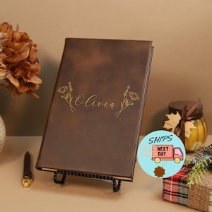 May include: A brown leather-bound journal with the name "Olivia" embossed in gold lettering. The journal is decorated with a gold floral design and is displayed on a black stand.